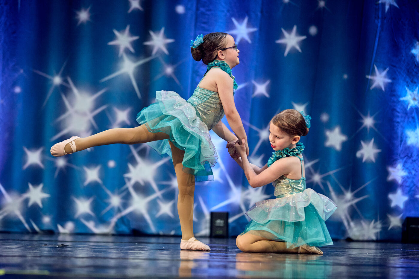 Two little ballerinas in teal tutus sharing a moment on stage
